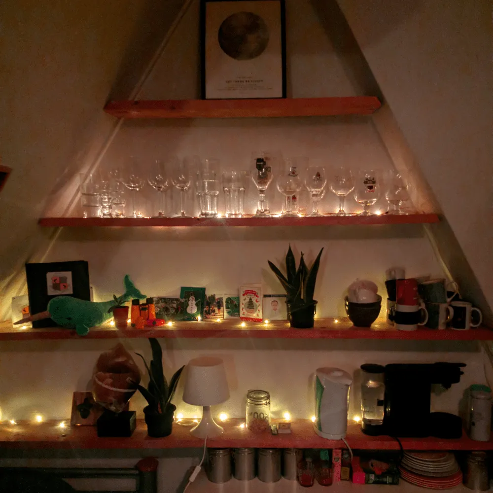 Triangular wall shelves under a sloped ceiling, each shelf lit by a string of warm fairy lights.